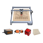 The Creality Falcon CR 5W Diode Laser Engraver showcases ultra-fine engraving on a wooden sheet with intricate patterns, displayed alongside orange safety glasses, a laser module, a red storage box, and extra wood sheets.