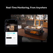A Creality Falcon A1 Pro 20W Dual-laser Engraver sits on a desk, while a smartphone displays real-time remote monitoring. Text above: "Real-Time Monitoring, From Anywhere." Includes free test kit.