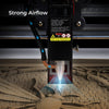 Close-up of a Creality Falcon Air Assist for CR-Laser Falcon 7.5W and 10W Engraver directing powerful airflow at a wooden surface. A bright laser beam is in action, surrounded by warning labels and specifications. Blue arrows show the airflow direction, maintaining low working noise while keeping material surfaces clean.