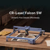 A 5W CR-Laser Falcon Engraver Upgrade Package by CrealityFalcon sits on a wooden table with engraved wood art beside it and the text "CrealityFalcon 5W CR-Laser Falcon – Achieve Fine Details Effortlessly" displayed above.