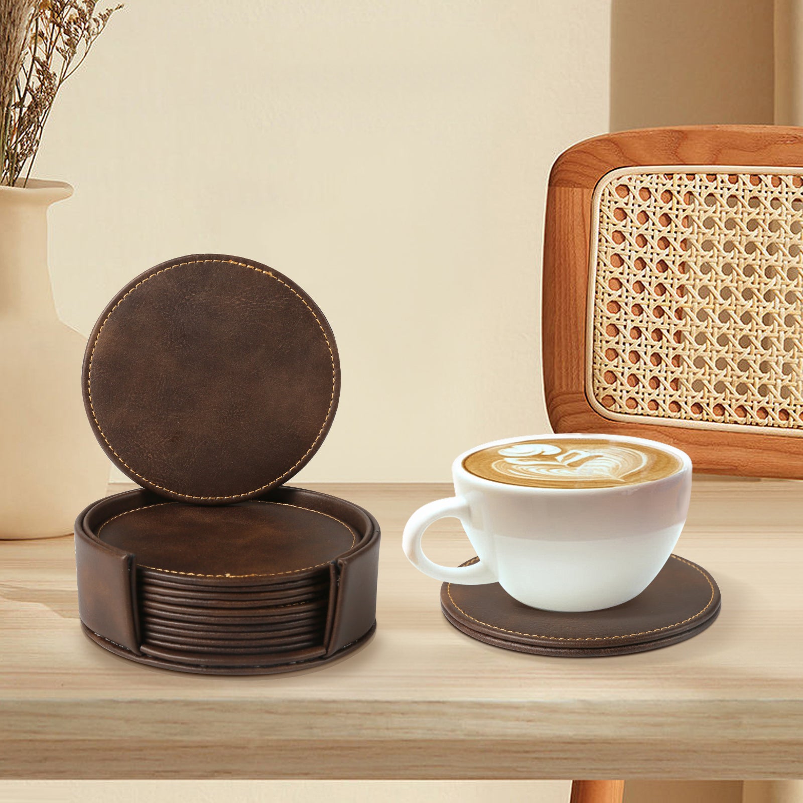 A coffee cup with latte art sits on a dark Creality Falcon coaster, showcasing modern design, atop a wooden table. A matching set of Personalized Brown Leather Coasters by Creality Falcon is stacked in a holder nearby. In the background are a chair with a woven backrest and a vase of wheat stalks.