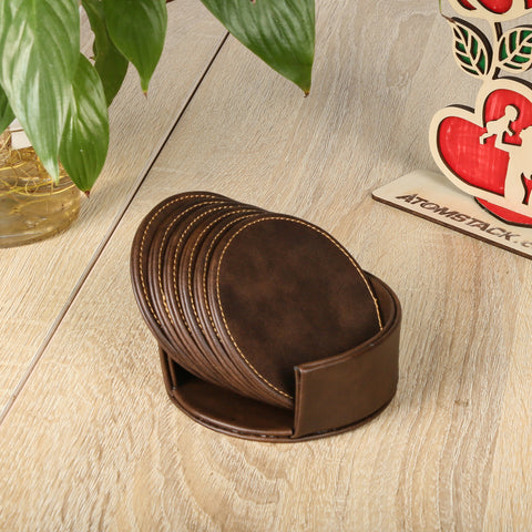 The Personalized Brown Leather Coasters and Holder by Creality Falcon, featuring white stitching and modern design, elegantly rest on a wooden table. Nearby, a green plant complements a heart-shaped decorative piece with cutout figures.