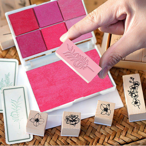 A hand holds a Creality Falcon wooden rubber stamp above a pink ink pad, from the Wooden Rubber Stamp Set with 6-Color Ink Pad for Crafting - 14pcs. Several other DIY floral stamps are scattered nearby on a wicker surface.