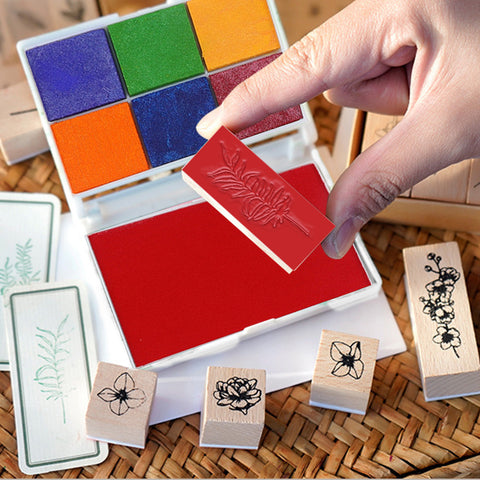 A hand holds a pink rubber stamp with a leaf pattern above a red ink pad. Nearby, Creality Falcon's Wooden Rubber Stamp Set with intricate floral designs and a 6-color ink pad sits on a woven surface.